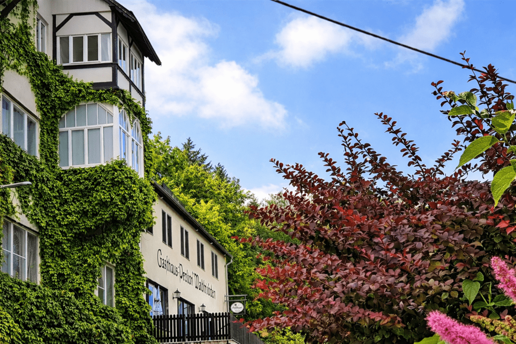 Gasthaus‑Pension Waldfrieden in Wolkenstein, umgeben von sommerlicher Natur – einladende Fassade mit warmem Licht und blühenden Sträuchern.